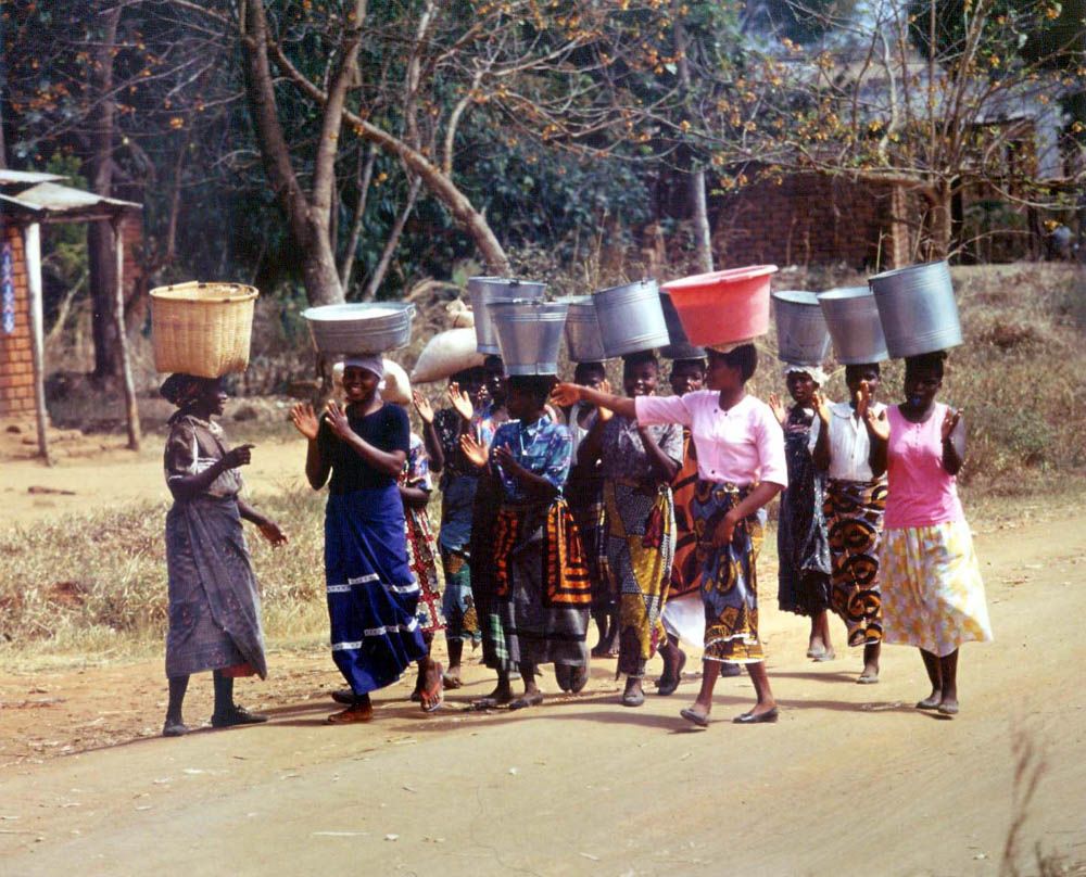 A line of women walk down a road, laughing, with metal buckets balanced on their heads.
