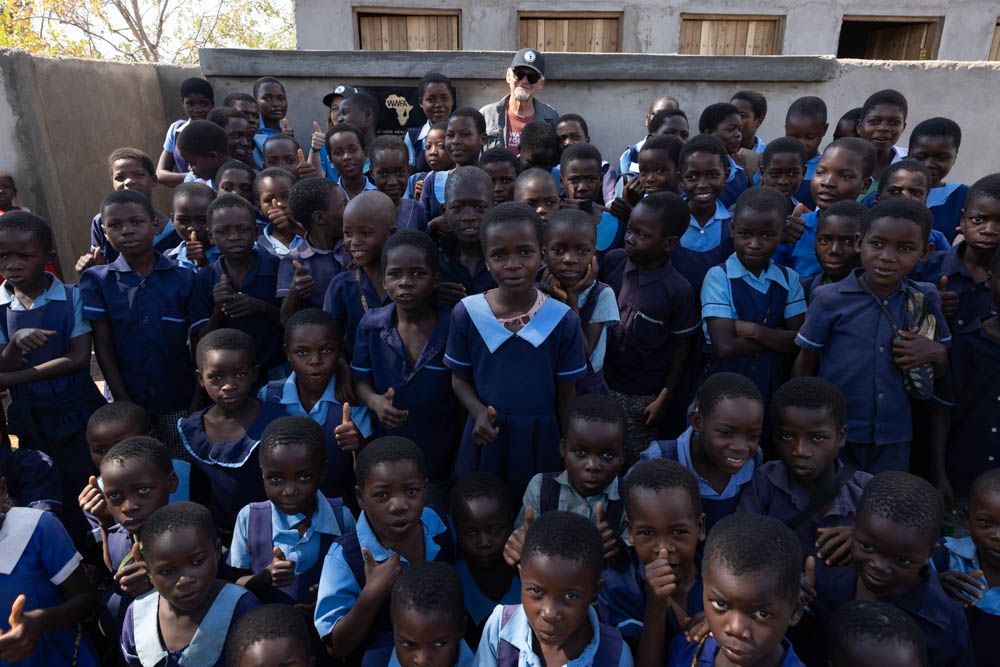 Dozens of schoolchildren in blue uniforms crowd together, many giving a thumbs up.