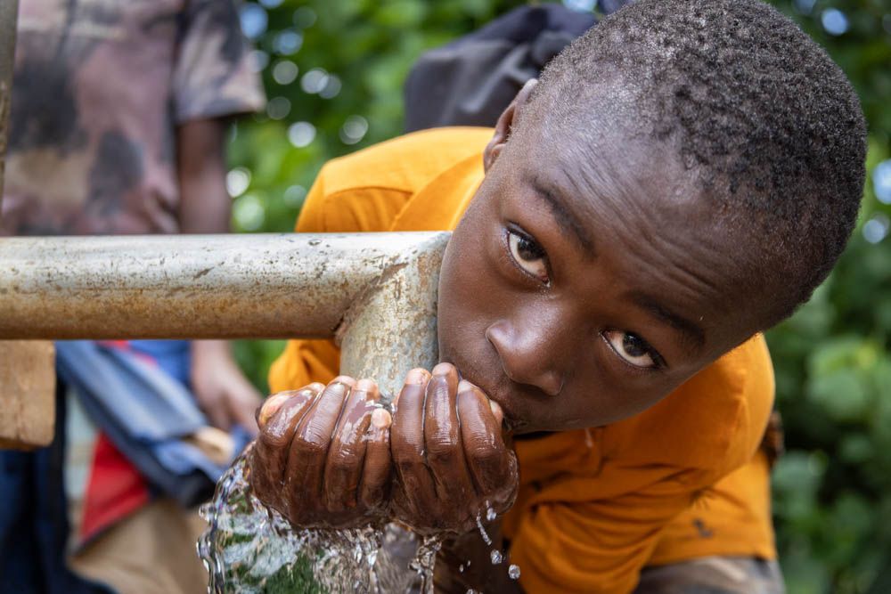 A boy in an orange shirt drinks straight from the spout of a hand pump.