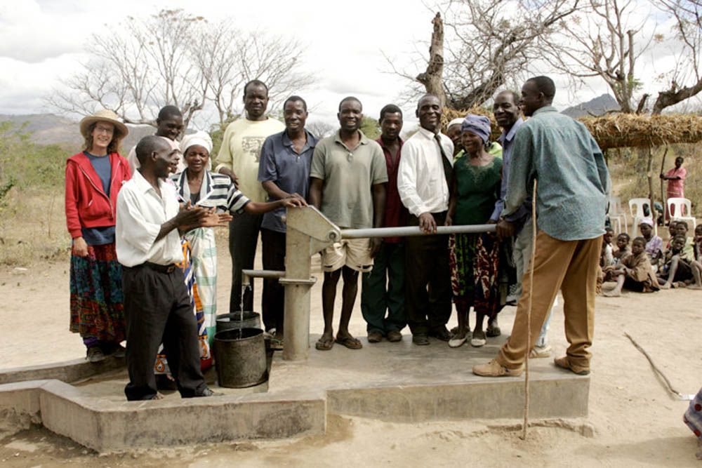 Villagers stand together around a hand pump as water pours into a bucket.