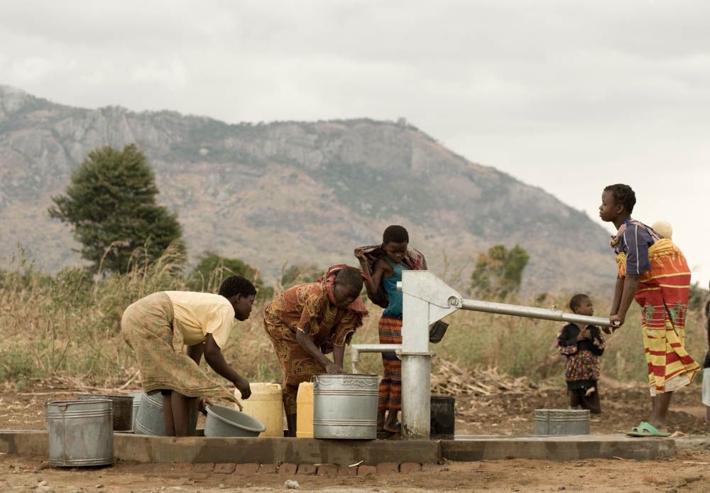 Women and children fill buckets at a hand pump below a mountain.