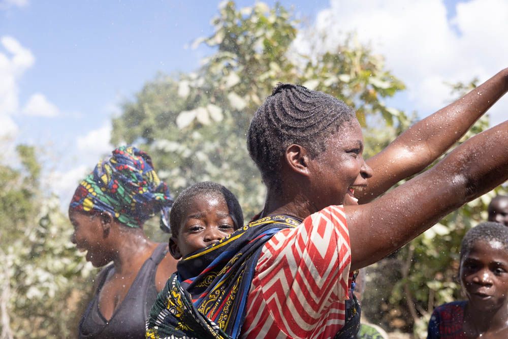A woman with a baby on her back laughs as water sprays around her.
