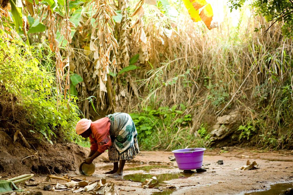 A woman bends to scoop water from a shallow puddle in a dry riverbed.