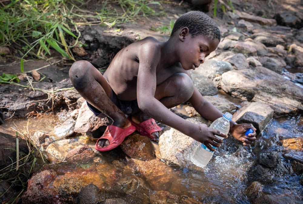 A boy crouches on rocks, filling a plastic bottle from a muddy stream.